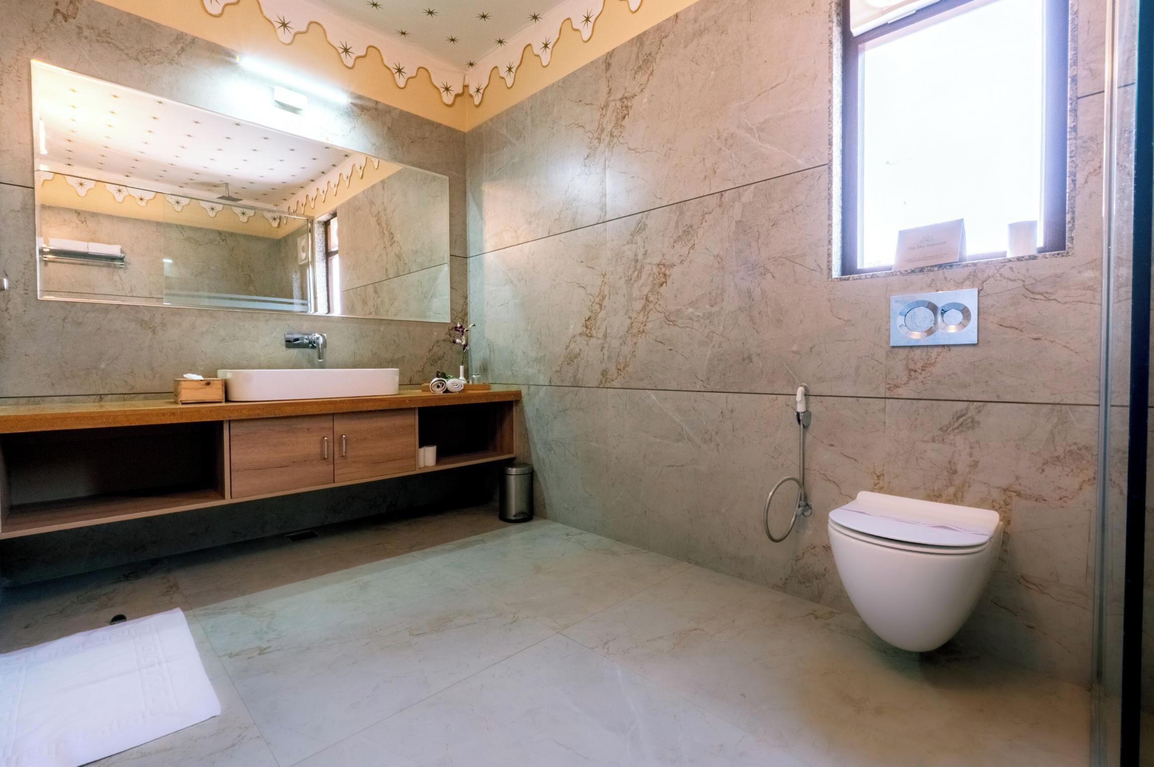 Un baño con pared de mármol, tocador de madera, lavamanos blanco, espejo, inodoro y ventana.