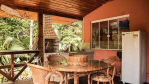 Uma área de refeições coberta ao ar livre, com mobiliário de vime, uma mesa de vidro e uma geladeira branca.