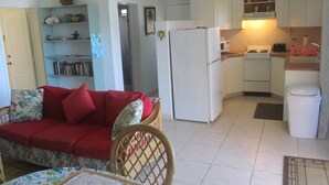 Un espacio habitable compacto con kitchenette, un sofá con cojines rojos, una mesa de comedor y una silla de mimbre.