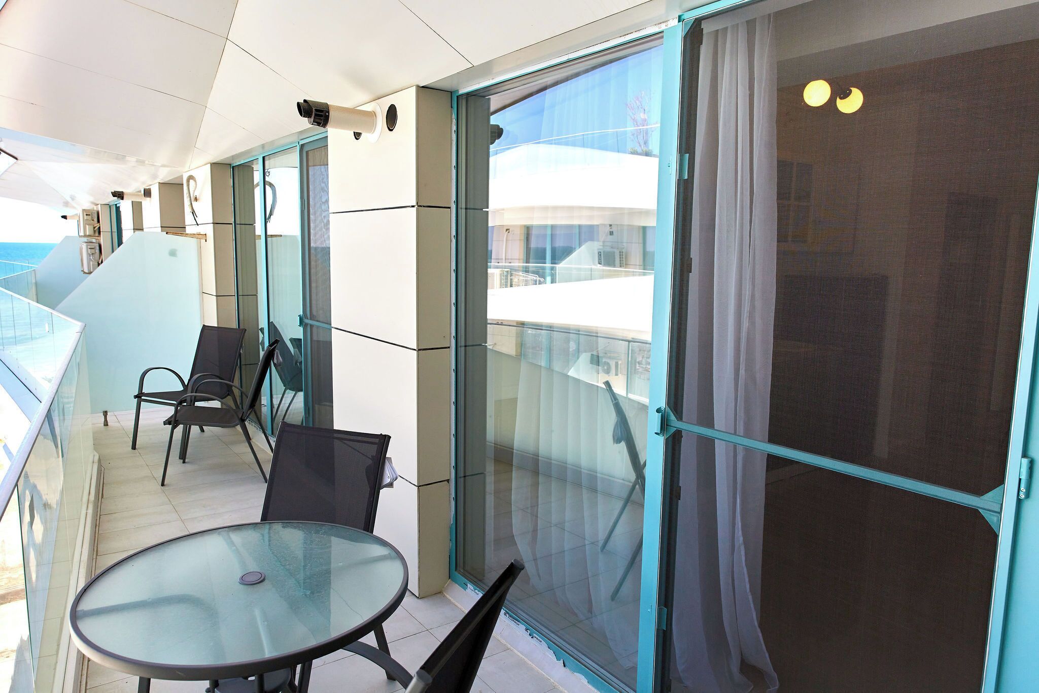 A balcony with a glass table and chairs, a sliding glass door, and a view of the sea.