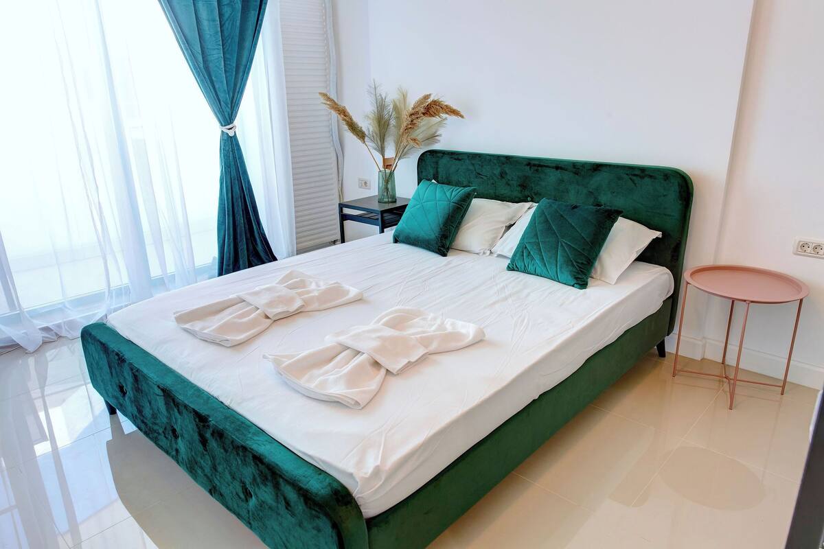 A neatly made bed with white linens and green pillows, a small bedside table with a vase of dried pampas grass, and a pink stool.
