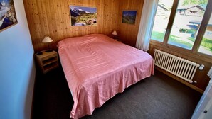 A bedroom with wooden walls, a bed with a pink bedspread, two bedside lamps, a window with curtains, and a radiator. - Pre Vert D62, Morgins (Morgins)