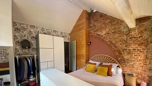 Ein Schlafzimmer mit Backsteinwand, Holzboden, einem Bett mit gelben Kissen, einem weißen Schminktisch und einem Holzschrank.