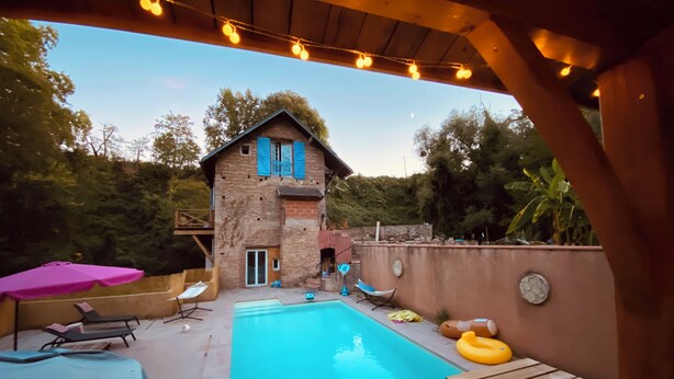 Ein Garten mit Schwimmbecken, einer Holzterrasse, Liegestühlen und einem Steinhaus mit blauen Fensterläden.