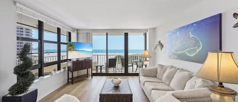 A modern living room with a large window overlooking a beach, a comfortable sofa, a coffee table, and a painting on the wall.