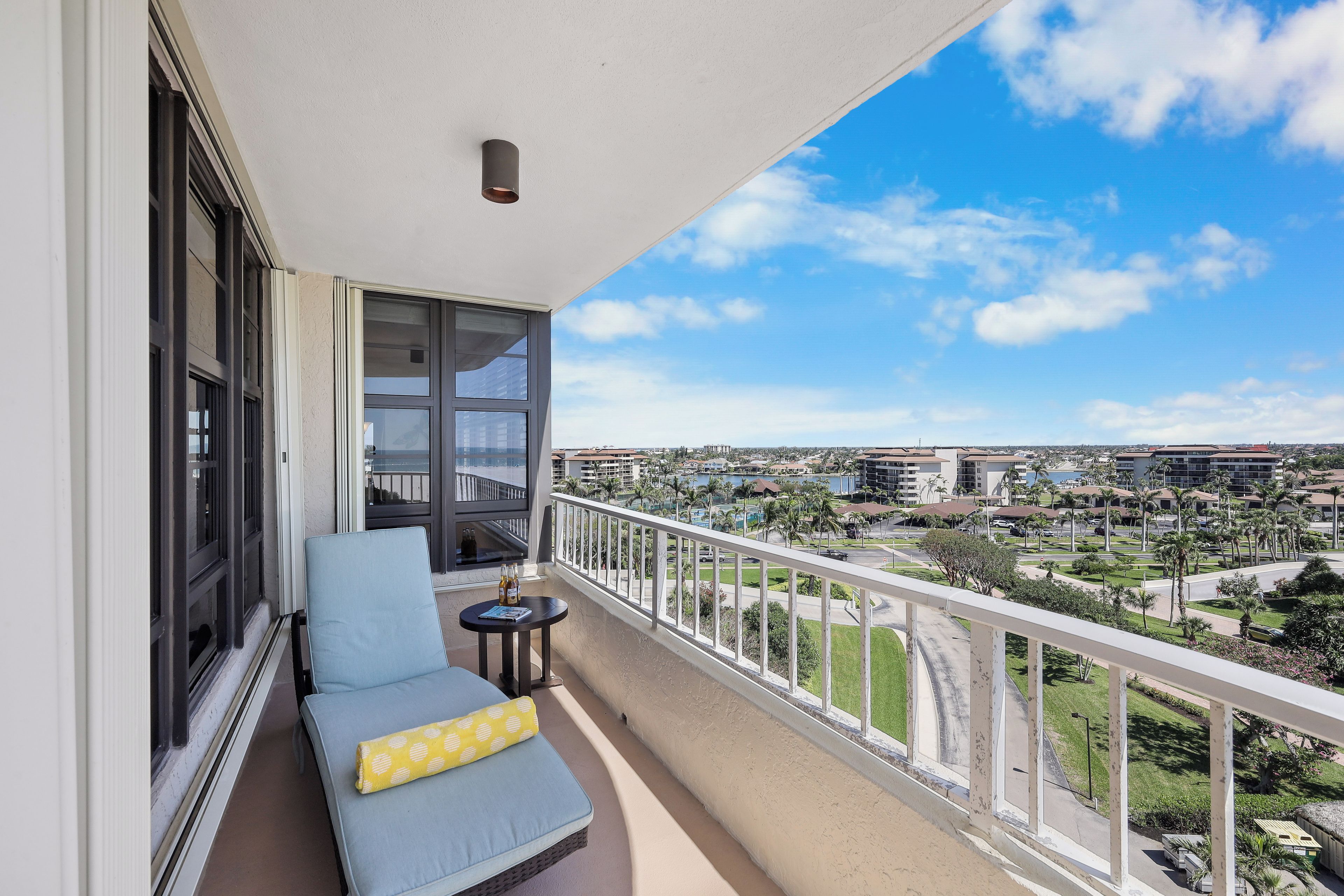 A balcony with a sofa and a small table, overlooking a cityscape with buildings and greenery.