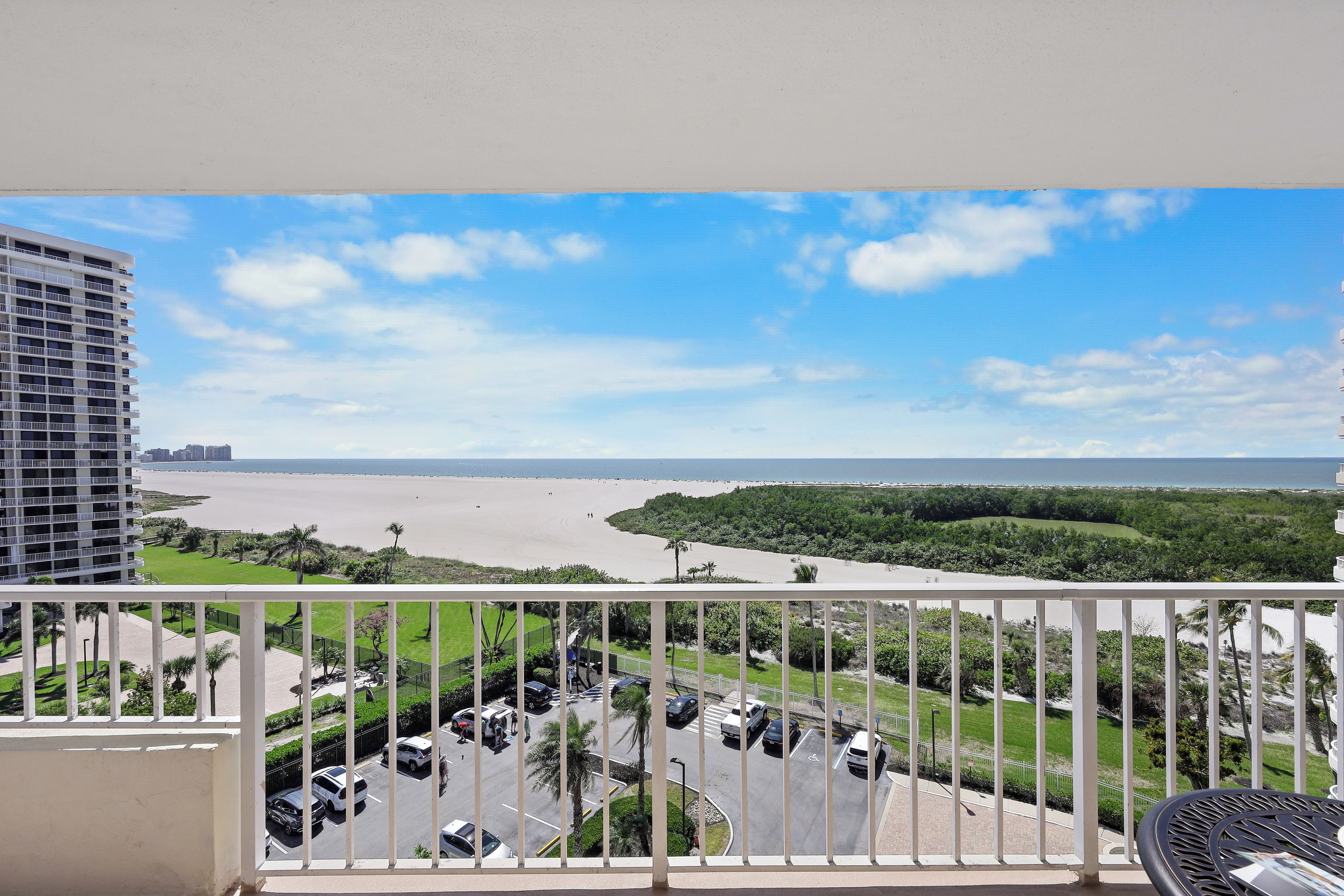 A balcony with a view of a beach, parking area, and greenery.