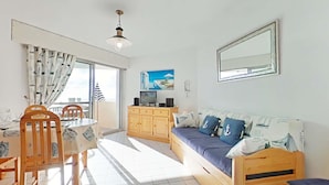A bright living room with a wooden sofa, a dining table with chairs, a TV, and a large window with curtains. - Studio 1 room, sleeps 4, Saint Gilles Croix de Vie, Parking (Saint-Gilles-Croix-de-Vie)