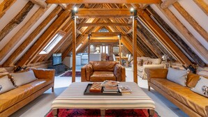 A cozy attic bedroom with a high ceiling, exposed wooden beams, a central bed with a red patterned rug, two leather sofas, and a small kitchenette in the background. - Belchamp Hall Hayloft and Stables, Belchamp Walter (Belchamp Walter)
