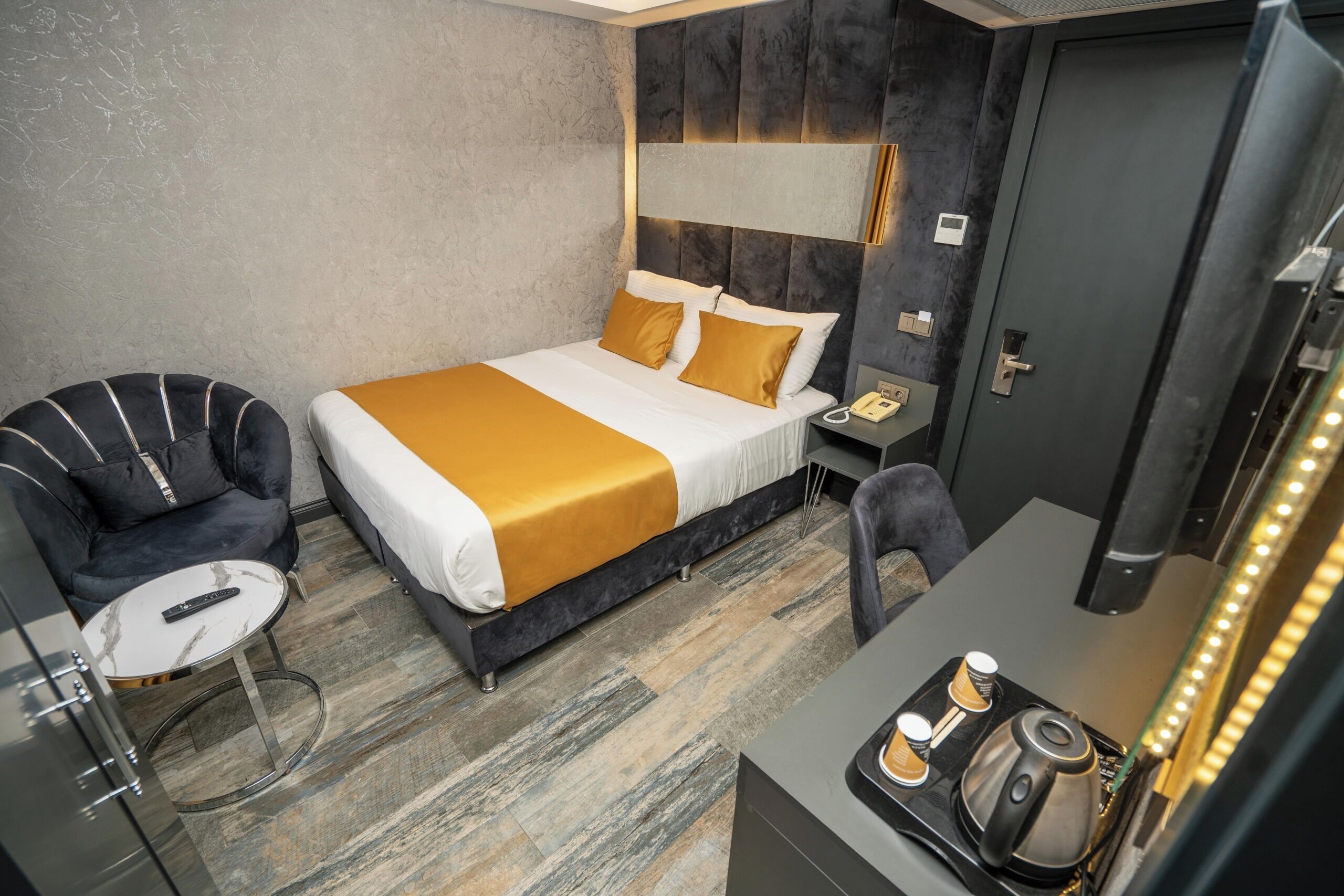 A modern hotel room with a large bed, a desk with a coffee machine, a chair, and a bedside table with a phone.