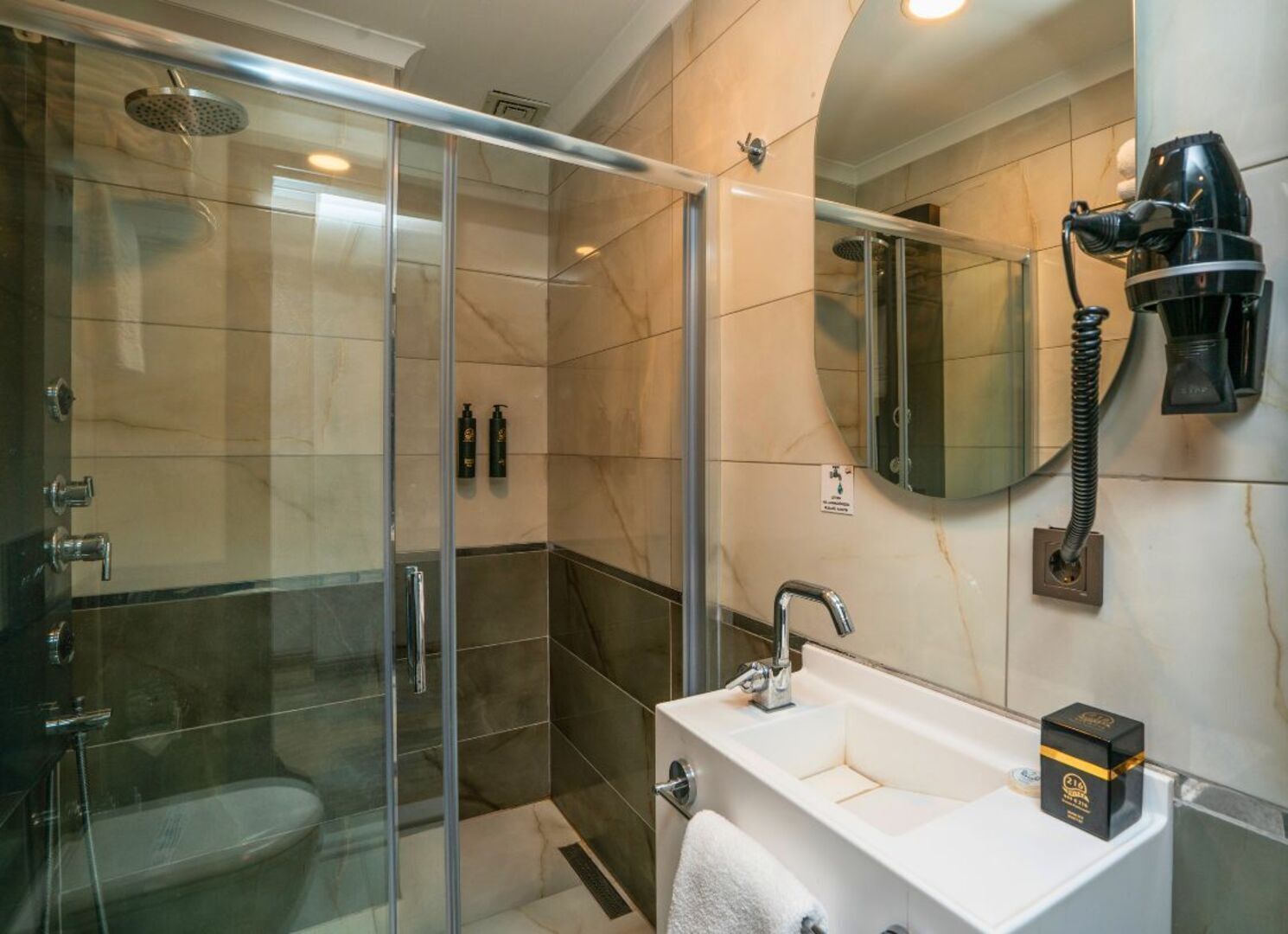 A modern bathroom with a glass-enclosed shower, a white sink, and a hairdryer mounted on the wall.