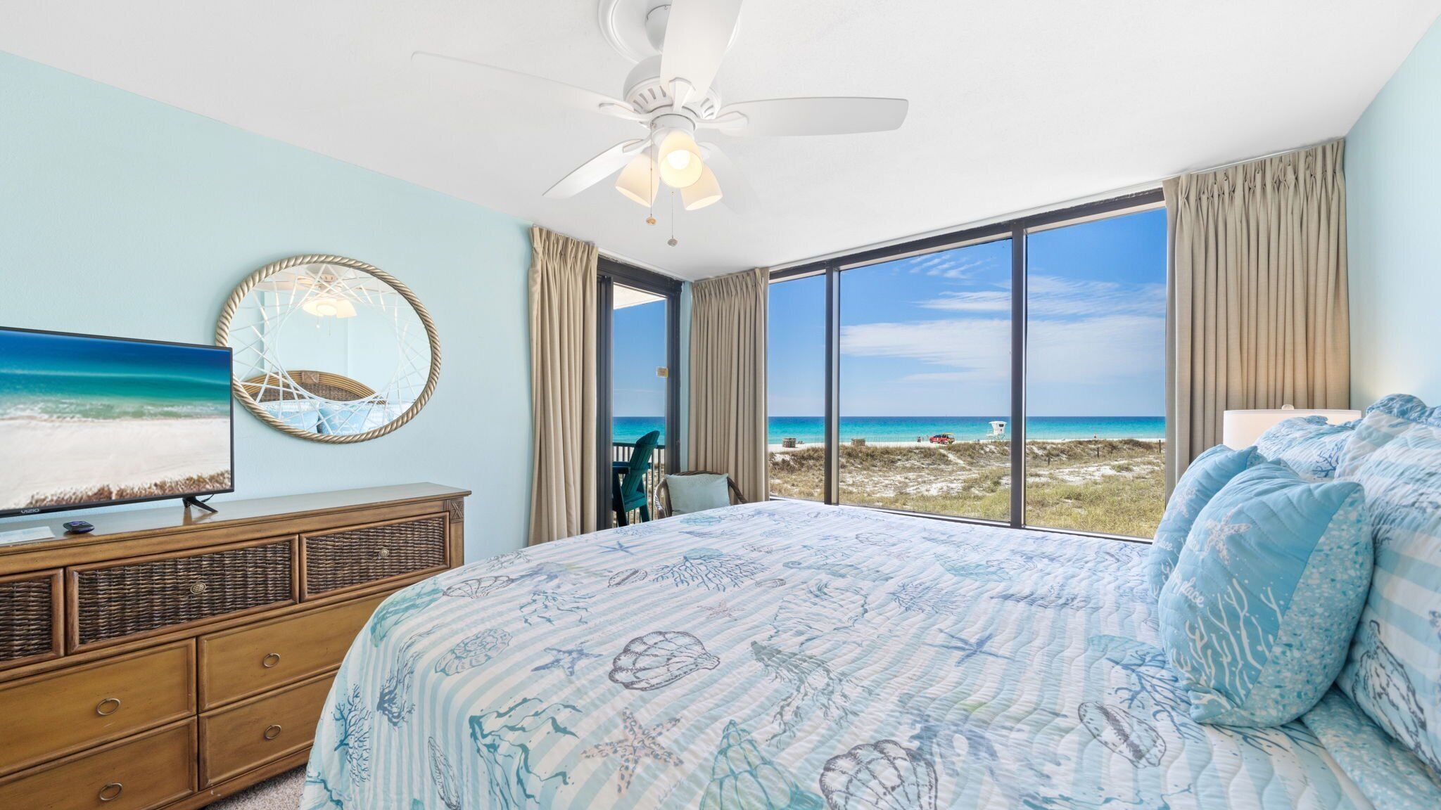 A bedroom with a large bed, a ceiling fan, a TV, and a view of the beach.