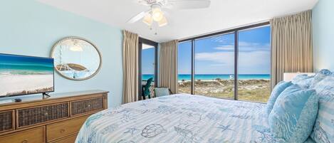 A bedroom with a large bed, a ceiling fan, a TV, and a view of the beach.