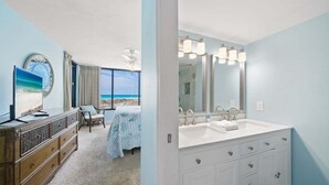 Habitación de hotel con un ventanal grande que ofrece vista a la playa, un lavamanos doble y una cama con vista, además de una silla junto a la ventana.