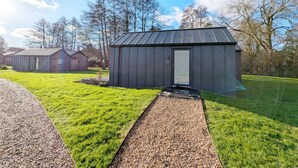 Un piccolo capanno di metallo scuro con una porta, circondato da un'area verde curata con un vialetto in ghiaia che vi conduce.