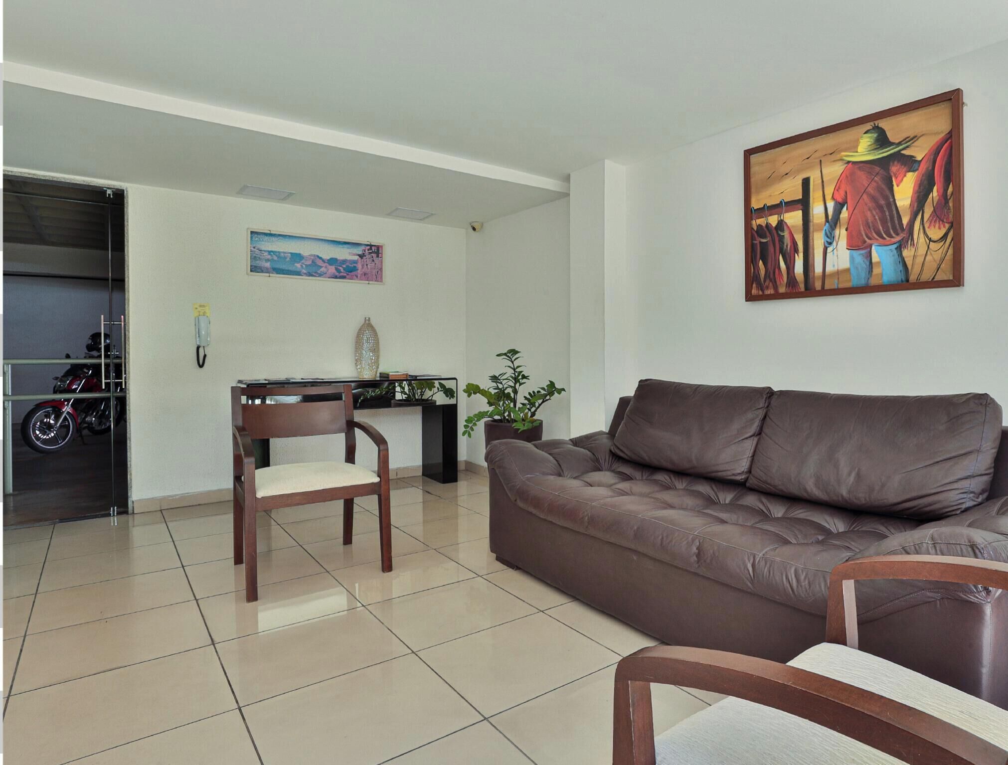 Una sala de estar con un sofá de cuero marrón, una silla de madera, una mesita, una planta en maceta y un cuadro en la pared.