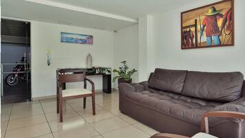 Una sala de estar con un sofá de cuero marrón, una silla de madera, una mesita, una planta en maceta y un cuadro en la pared.