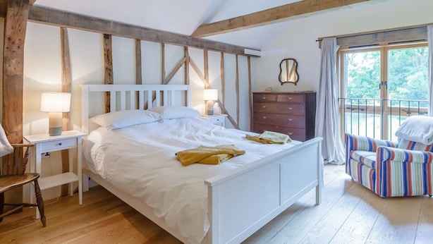 Une chambre avec un plafond en bois, un lit avec une literie blanche, une commode, une chaise et une fenêtre avec des rideaux.