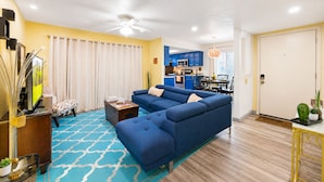 A living room with a sectional sofa, a television, a coffee table, and a dining area with blue cabinets and a kitchen in the background.