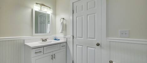 Un baño con tocador blanco, espejo con iluminación, una puerta y un perchero para toallas.