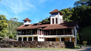 A large, two-story building with a red-tiled roof and multiple windows, surrounded by lush greenery and a clear blue sky. - Sana Spa Maria Bonita (Macaé)