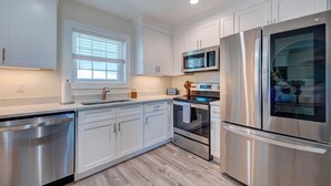 A modern kitchen with white cabinets, stainless steel appliances, and a window with blinds. - 2005 Queen Suite, 2nd Floor Surfside (Surf City)