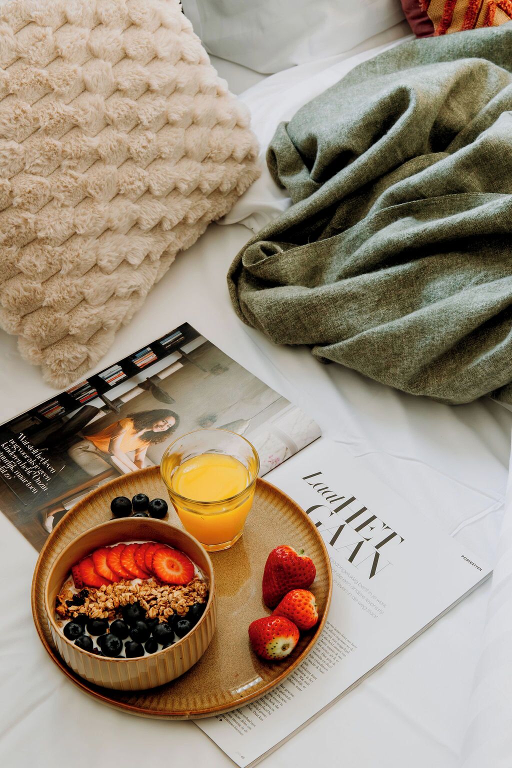 A neatly arranged breakfast setting with a bowl of yogurt topped with granola, strawberries, and blueberries, a glass of orange juice, and a magazine on a bed.