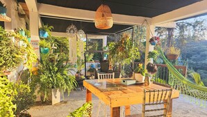 Un patio couvert avec une table et des chaises en bois, entouré de différentes plantes en pot et d’un hamac.