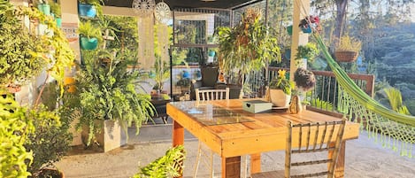 A covered patio area with a wooden table and chairs, surrounded by various potted plants and a hammock.