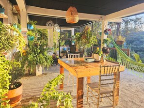A covered patio area with a wooden table and chairs, surrounded by various potted plants and a hammock. - Ecohotel Zona 7 (Rionegro)