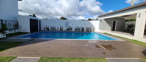 Una piscina con borde de baldosas, rodeada por un muro blanco y un pequeño jardín con césped y plantas.