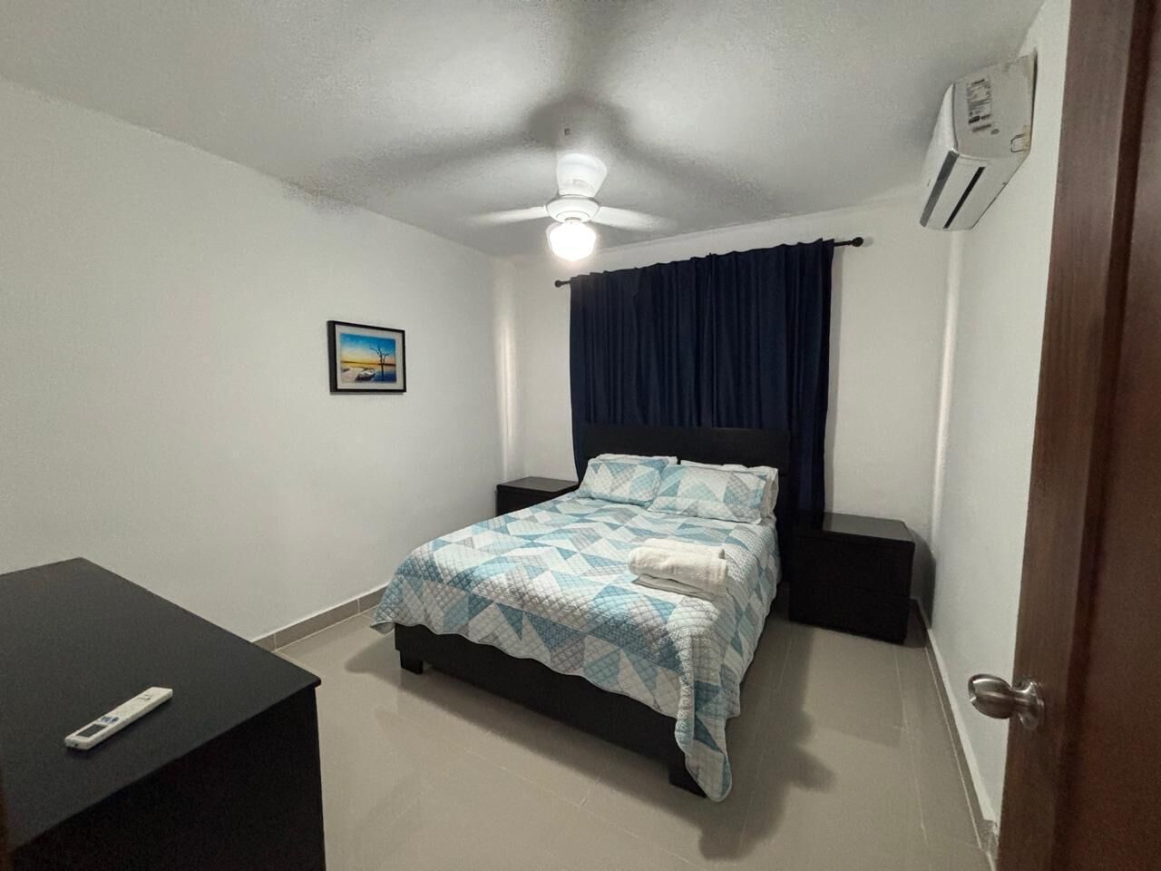 A bedroom with a bed, a desk, a ceiling fan, an air conditioner, and a picture on the wall.