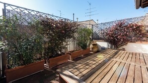 Een houten terras met plantenbakken en een bank, omringd door een metalen hek en beplanting.