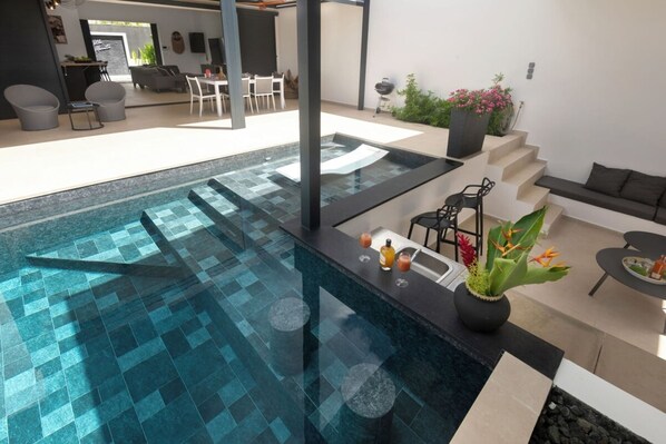 A modern indoor pool area with a bar, seating, and a staircase leading to an upper level.