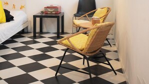 Een kleine, schone kamer met een geruit vloerkleed, een bed met gele kussens, twee rieten stoelen, een klein tafeltje met een radio, en een hanglamp.