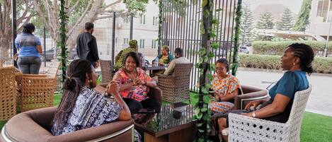 Un grupo de personas sentadas en un café al aire libre con muebles de mimbre y una mesa de vidrio.