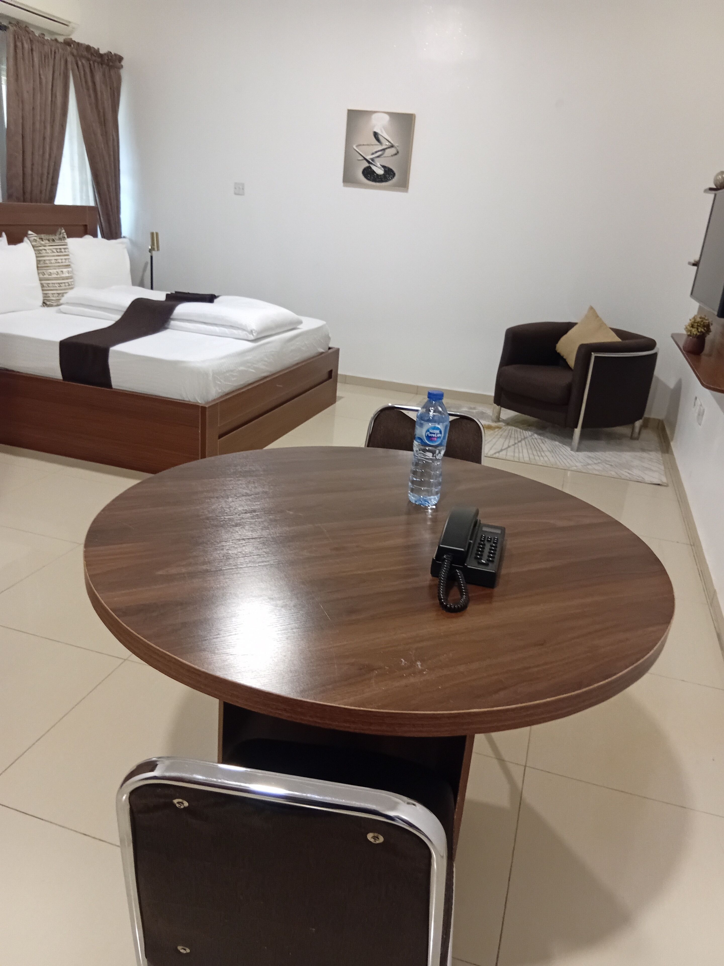 A hotel room with a bed, a round dining table with chairs, a water bottle, a telephone, and a small side table with a lamp.