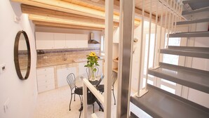 Une cuisine moderne avec des armoires blanches, un pare-feu en carrelage et un plafond en bois. On y trouve un coin repas avec une table et des chaises, ainsi qu’un escalier doté d’une rampe en métal.