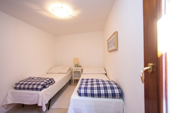 A small, well-lit room with two single beds, each with a plaid blanket and pillow. There is a small table with a lamp and a framed picture on the wall.