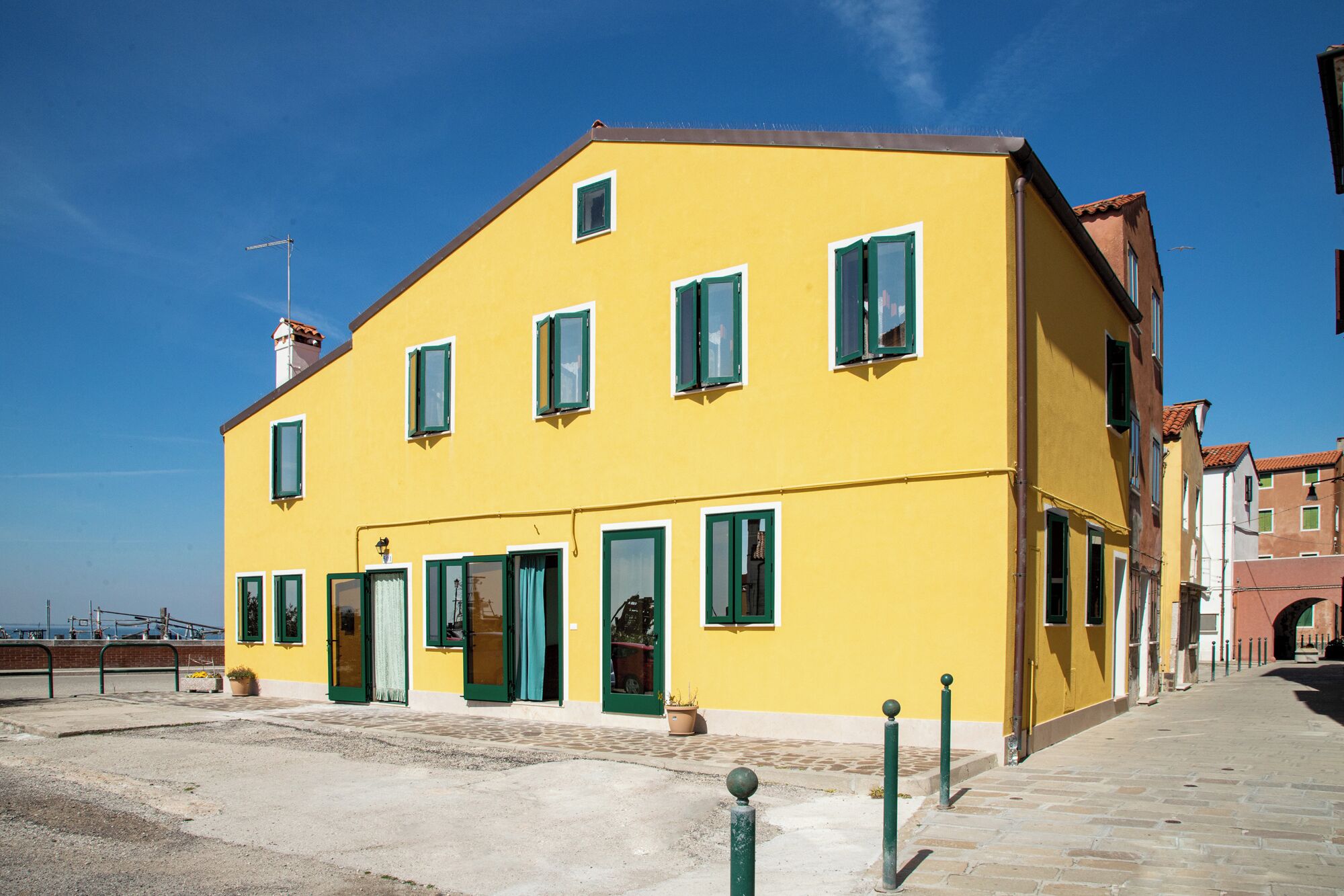 A yellow building with green shutters, a visible entrance, and a paved area in front.