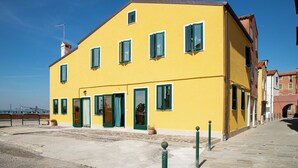 A yellow building with green shutters, a visible entrance, and a paved area in front. - Dimora Stella  -apeliving- Pellestrina Ve (Venice)
