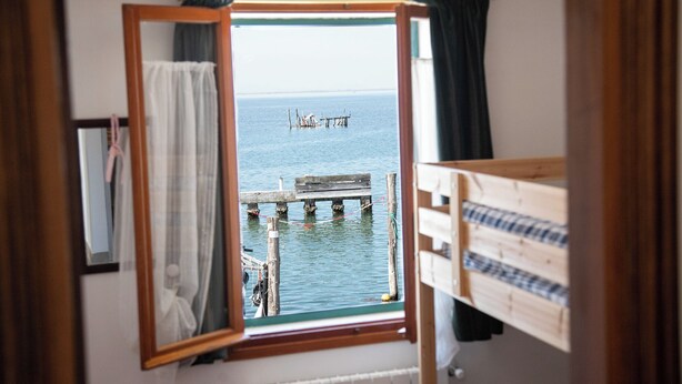 A room with a wooden bunk bed, a window with a view of a dock and water, and a curtain. - Dimora Stella  -apeliving- Pellestrina Ve (Venice)