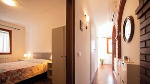 A bedroom with a bed, bedside lamps, and a wardrobe. There is a hallway with a potted plant and a mirror on the wall.