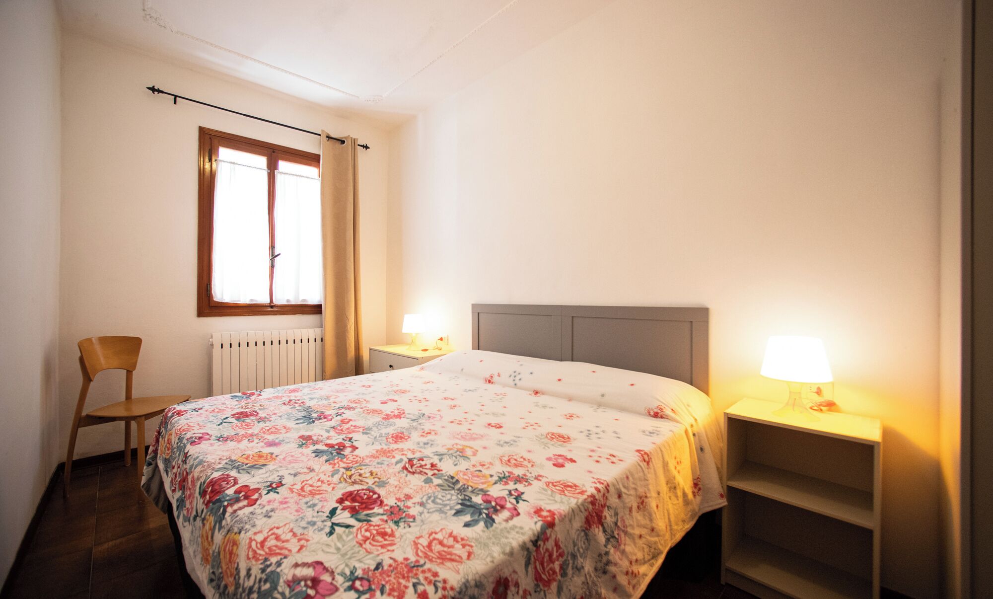 Een slaapkamer met een bed, twee bedlampen, een stoel, een raam met gordijnen en een radiator.