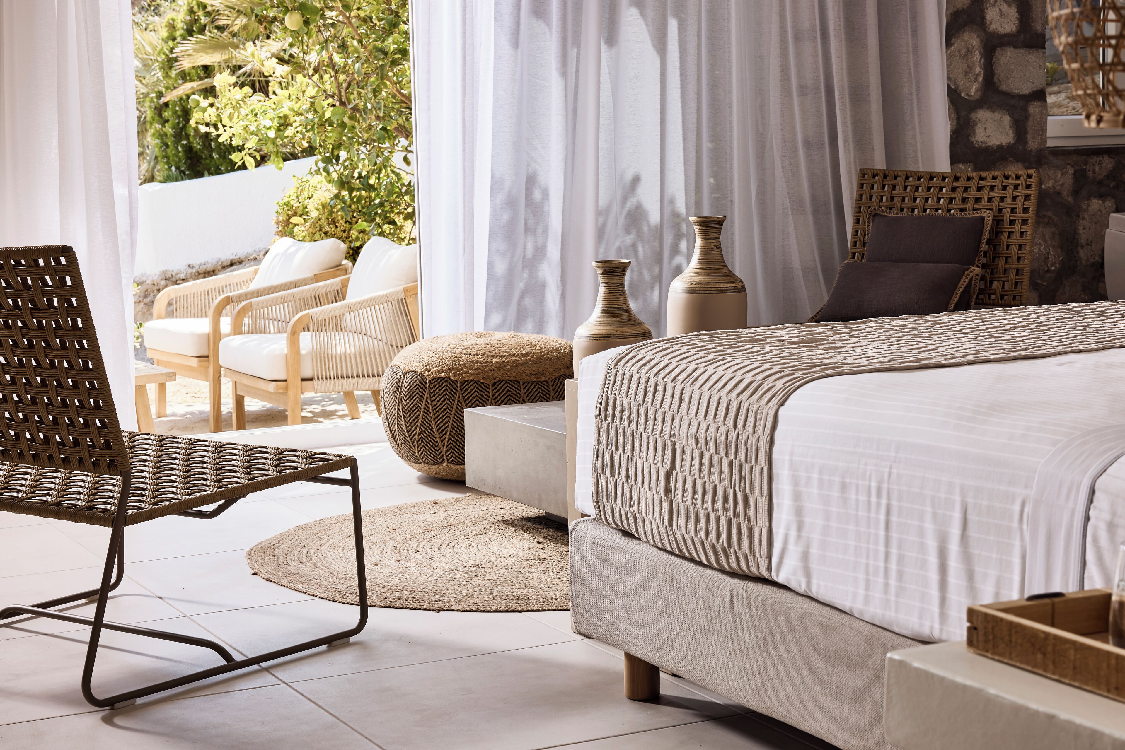 A bedroom with a bed, a wicker chair, a round ottoman, and a view of greenery outside.