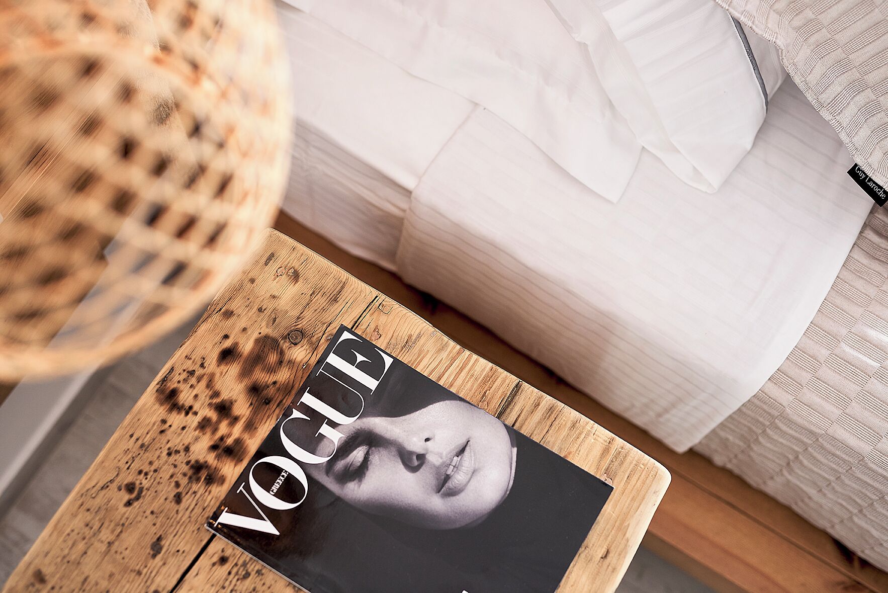 Una revista sobre una mesa de madera, una cama blanca y una cesta tejida.