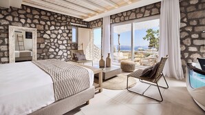 Camera da letto con parete in pietra, un letto grande, una sedia, un tavolino e vista sull'esterno.