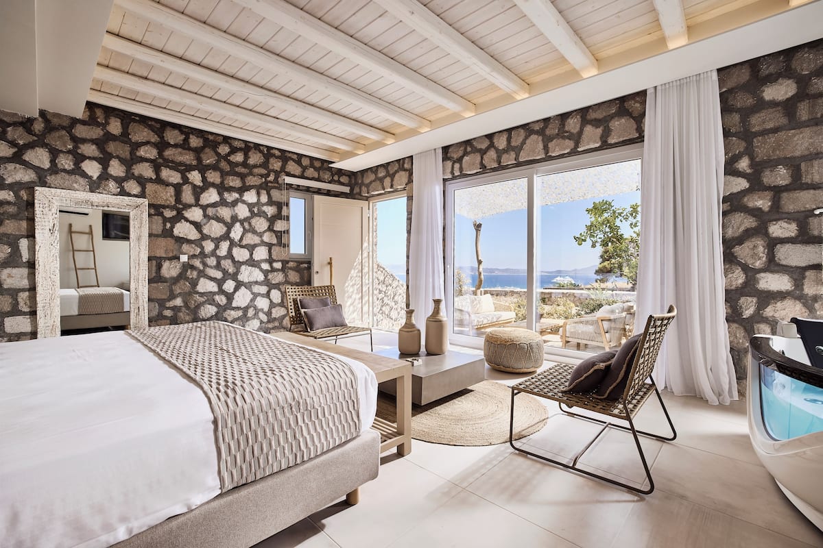 Une chambre avec un mur en pierre, un grand lit, une chaise, une petite table et une vue sur l’extérieur.