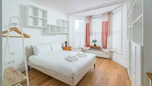 A bedroom with a bed, a radiator, a window with curtains, a wooden floor, and white walls. - Stunning house near Notting Hill (London)