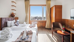 Una camera d'albergo con due letti, una testiera in legno, una scrivania con sedia, una finestra con tende e vista sulla città.